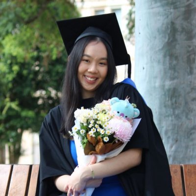 A young woman in graduation cap and gown sits on a bench seat in 喵喵直播's Great Court, clutching a bouquet of flowers with a small teddy bear inside it.